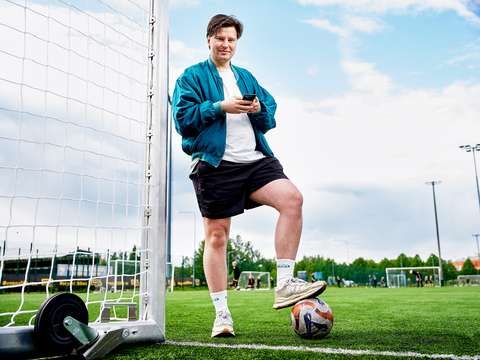 Elias Reunamo is standing on a football field next to the goal. Reunamo is looking at the camera, holding a smartphone in his hands. He has one foot resting on a football.