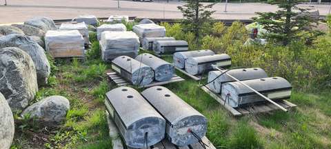 Kymmenen graniittivalaisinta puulavojen päällä nurmikolla. Taustalla kelmuun pakattuja puulaatikoita.