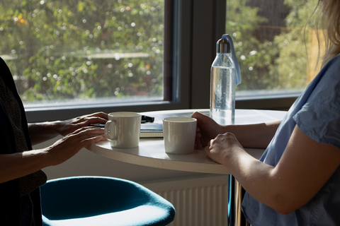 Kaksi henkilöä seisoo pystypöydän ääressä, toinen pitää kädessään kahvikuppia ja toisen kämmenet nojaavat pöytään.