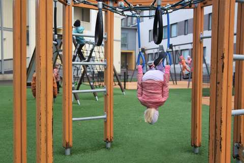 In the picture a child is hanging up side down on a climbing frame. In the back groung there are other children playing and a white building surrounds the area.