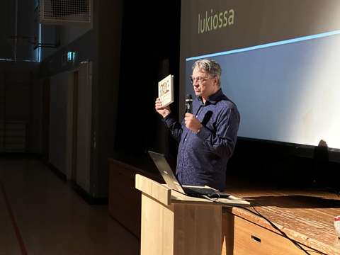 luennoitsija juhlasalissa kirja kädessä