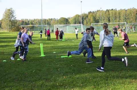Kuvassa ryhmä koululaisia juoksee nurmikentällä eri suuntiin. Maassa on kirkkaan vihreä kritettimaila.