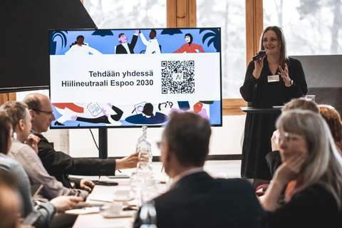 Lavalla puhuja kustuu yityksiä mukaan Hiilineutraali Espoo 2030 sitoumukseen. Puhujan vieressä näyttö jossa QR koodi.