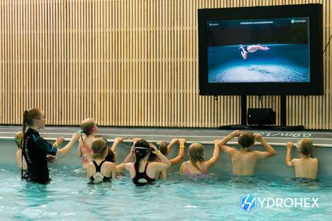 Barn vid kanten av simundervisningspoolen som tittar på instruktionsvideor på en digital skärm.