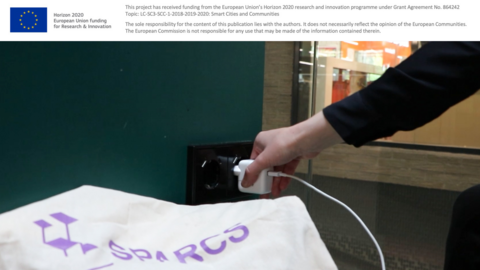 Kuvituskuva, jossa puhelimen laturia laitetaan pistorasiaan. Kuvan etualalla on kangaskassi, jossa on SPARCS-hankkeen logo.