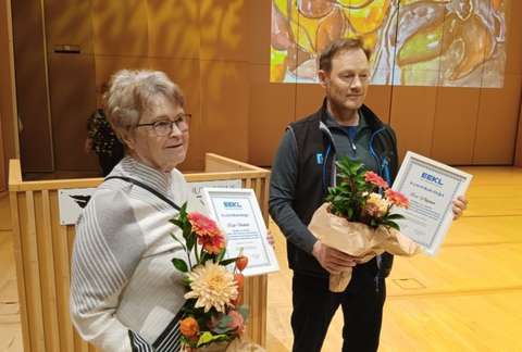 Vanhempi nainen ja keski-ikäinen mies seisovat lavalla kunniakirjat ja kukat käsissään.