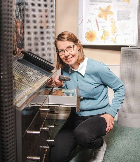 A smiling Katri Luukkonen standing in front of a display case with a magnifying glass in her hand and looking at the camera.
