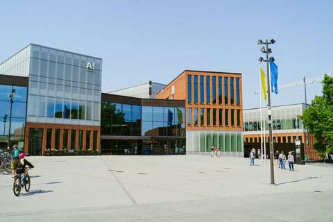 A building with a square in front of it. There is a cyclist and pedestrians in the square.