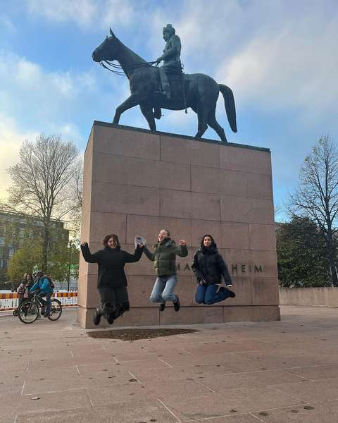 Kolme tyttöä hyppää patsaan edessä ilmaan.