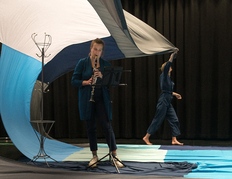 Henkilö etualalla soitta instrumenttia ja taustalla toinen henkilö nostaa kangasta ilmaan.