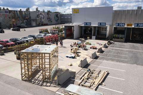 I förgrunden en kubikformad konstruktion av trä och i bakgrunden logistikhallarna.