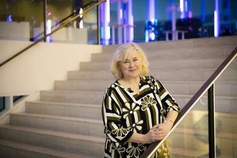 Leena Hoppania seisoo portaikossa ja nojaa kaiteeseen. Hänen takanaan näkyy isojen ikkunoiden takaa heijastuvaa sinistä valoa.