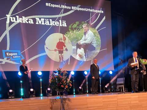 Kuvassa pukuun pukeutunut mies puhuu mikrofoniin lavalla. Lavan valkokankaalle on heijastettu kaksi kuvaa ja teksti Espoon Urheilugaala.
