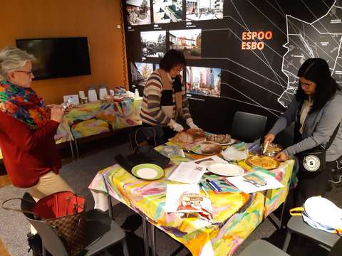 Kolme ihmistä valmistelevat pöydän äärellä leipäiltaa asettamalla erilaisia leipiä viipaloituina esille.