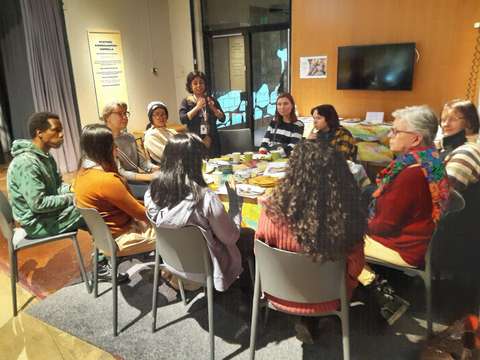 Pöydän ympärillä istuu kymmen ihmistä, yksi seisoo ja puhuu. Pöydällä on tarjolla erilaisia leipiä.