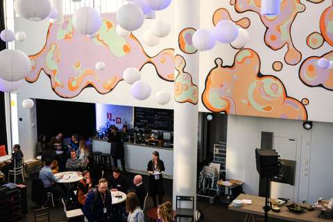 An overview of an event venue. The venue has a mural in pastel tones and there are several people are talking in the room.