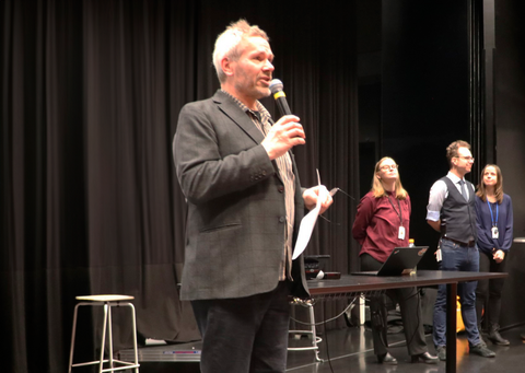 Mies puhuu mikrofoniin. Taustalla ihmisiä.