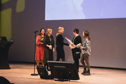 Viisi henkilöä esiintymislavalla. Kolme on luovuttamassa palkintoa ja kaksi vastaanottamassa.