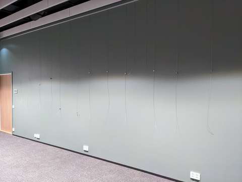 Tyhjä, harmaaksi maalattu näyttelyseinä, jota vasten roikkuu taulujen ripustusvaijereita.