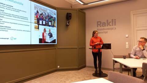 En kvinna klädd i en röd blus står bredvid en stor skärm i en seminarielokal och håller ett föredrag.