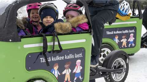 Muksubussissa istuvat lapset katsovat hymyillen kameraan, muksubussin kyljessä teksti: tässä kulkee kallis lasti.