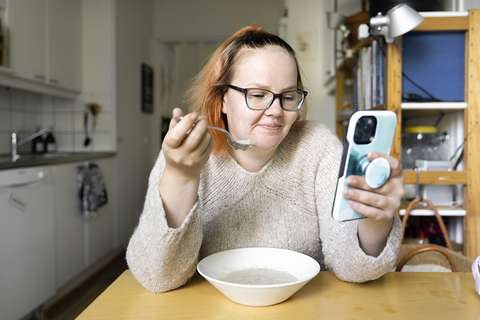 A person sits at a table eating porridge and reading a book on their phone.