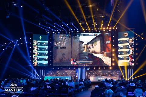 Kuvassa näkyy pimeässä tilassa videopeliä isolla näytöllä. Etualalla näkyy ihmisiä istumassa katsomossa.