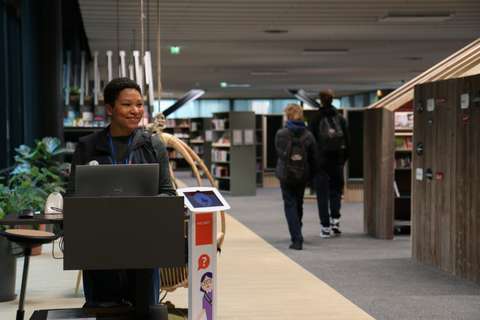 Lippulaivan kirjaston aulaa. Virkailija päivystää kevyen, liikutettavan tiskin takana hymyillen. Takana näkyy kirjaston hyllyjä ja kaksi asiakasta.