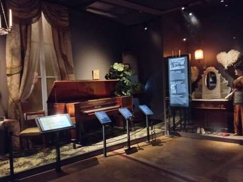 KAMUn näyttelyssä on esillä olohuonemaisessa tilassa piano, jonka edessä on neljä kylttiä. Kylteissä on näyttelytekstejä ja kuulokkeet. Kuvan oikeassa reunassa näkyy näyttelytekstin lisäksi vitriinissä oleva kananhöyhen puuhka, peilipöytä ja kolme paria kenkiä.