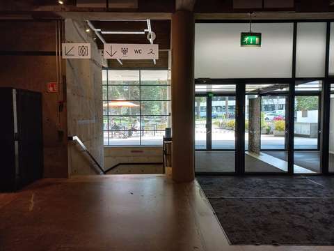 Kuvan vasemmalla puolella on WeeGeen sisällä aulassa portaiden yläpää ja kyltiti hissi, WC ja naulakko alakerran suuntaan. Kuvan oikealla on harmaa matto, jonka takana on WeeGeen lasiset sisäänkäynnin ovet.