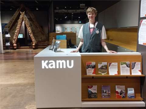 KAMUn näyttelytilassa harmaa tiski, jonka takana seisoo KAMUn museo-opas valmiina auttamaan ja kertomaan näyttelyistä lisää.  