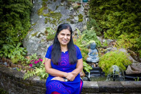 Tummatukkainen nainen istuu kivipenkillä ja hymyilee ystävällisesti. 