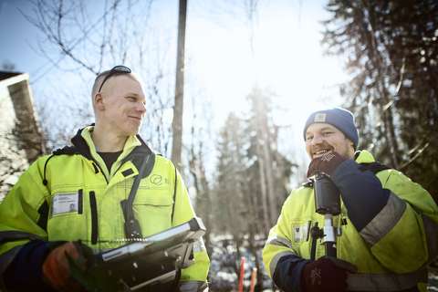 Two men in high visibility coats hold devices and smile in the winter sun.