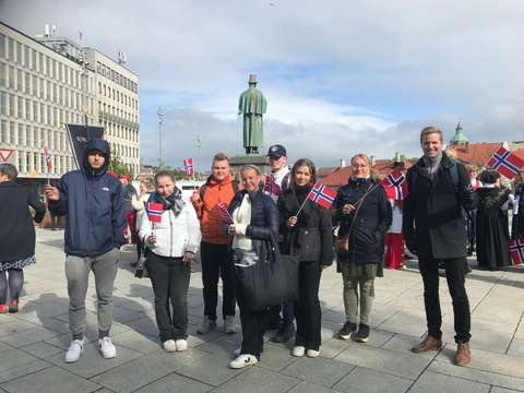 Opiskelijat ja opettajat seisovat patsaan edessä Norjan liput kädessään.