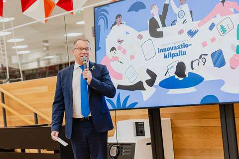 Mies puhuu mikrofoniin ja hänen taustallaan näkyy kuva, jossa lukee kaupunginjohtajan innovaatiokilpailu.