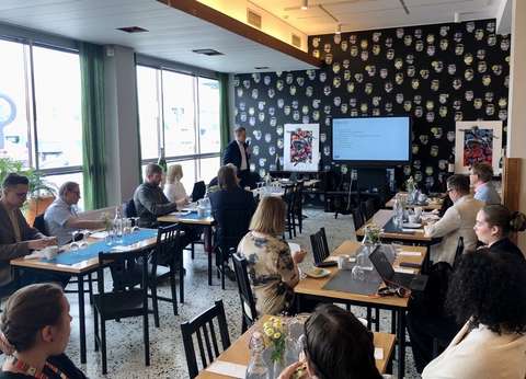 Ihmisiä pöytien äärellä kuuntelemassa tilaisuuden avauspuheenvuoroa. Etualalla puhuja ja näyttö.