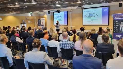 Yleisöä kuvattuna takaapäin istumassa aamiaisklubissa. Tapahtuman juontaja Saija Äikäs puhumassa.