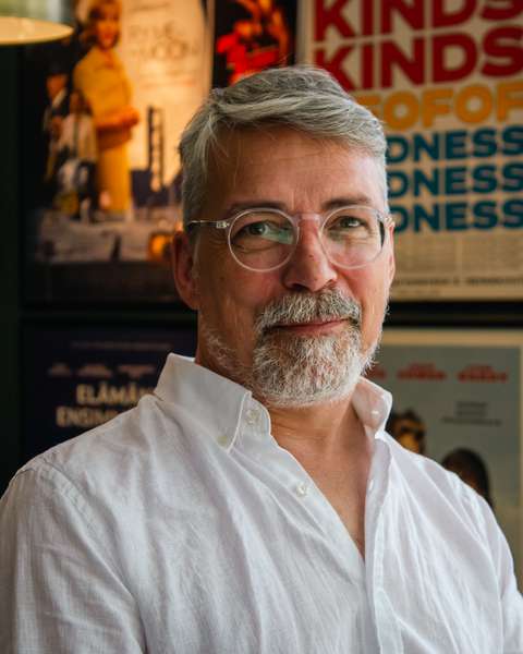 Parrakas vaaleihoinen mies, jolla on päällään valkoinen kauluspaita katsoo ystävällisesti kohti. Taustalla elokuvajuliste.l