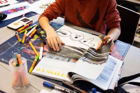 Nuori piirtää ja värittää graffitityötä paperille ja selaa samaan aikaa aikakauslehteä piirustusvälineiden ympäröimänä. 