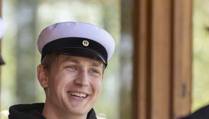 Kuvassa kolme nuorta opiskelijaa on ylioppilaslakit päässä. Yksi katsoo kameraan ja kaksi on selin.