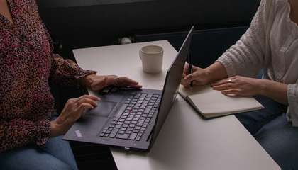 Two people sit at a table facing each other. The person on the left has a laptop open in front of them and the person on the right is taking notes.
