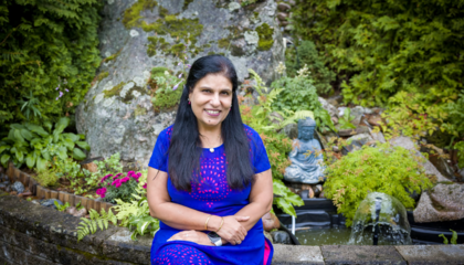 Tummatukkainen nainen istuu kivipenkillä ja hymyilee ystävällisesti. 