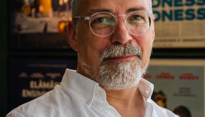 Parrakas vaaleihoinen mies, jolla on päällään valkoinen kauluspaita katsoo ystävällisesti kohti. Taustalla elokuvajuliste.l