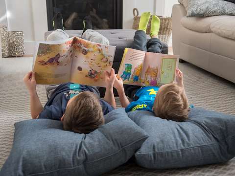 Two children are reading a book, lying on cushions on the floor.
