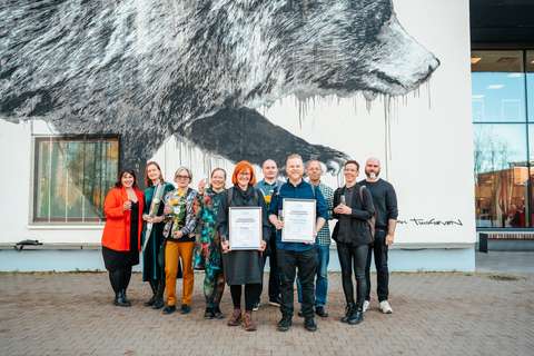 Ryhmä iloisia ihmisiä kohottaa maljoja ja esittelee kahta kunniakirjaa. Hei seisovat ulkona karhua kuvaavan muraalin edessä. 