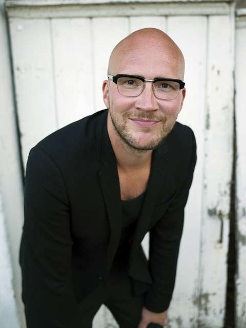 Mustiin pukeutunut kalju mies nojaa käsillä reisiin ja katsoo hymyillen kohti.