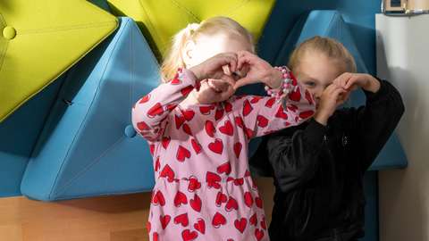 Kaksi päiväkoti-ikäistä lasta istuu vierekkäin ja tekee käsillään sydämet.