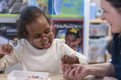 The child is making bead crafts and an employee is supporting nearby.