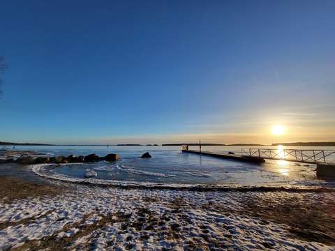Kuvassa näkyy laituri, jonka takana näkyy auringonlasku. Etualalla on rannan hiekkaa ohuen lumipeitteen alla ja veden pinnalla on jäätä.