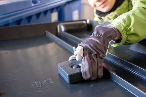 A person is placing a black, square-shaped sensor on the lid of a bin.